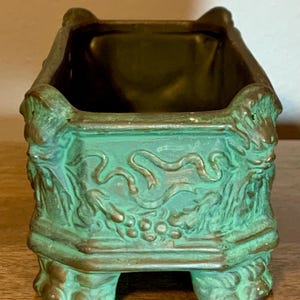 May include: A rectangular, antique-style planter with a verdigris finish. The planter features ornate relief detailing of vines, grapes, and ribbons. It has four decorative feet and a dark interior.