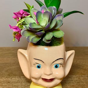 May include: A whimsical ceramic planter shaped like a smiling face with blue eyes, large ears, and a yellow collar. The planter is filled with a faux succulent, pink flowers, and green foliage, creating a unique decorative piece.