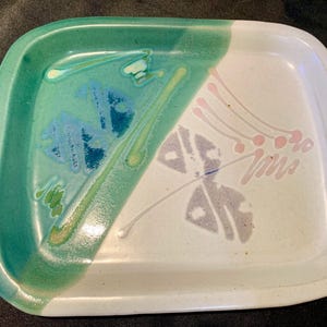 May include: A rectangular ceramic tray with rounded corners featuring a diagonal split design. One side is a vibrant green with blue and yellow abstract brushstrokes, while the other side is off-white with delicate pink and grey painted accents.