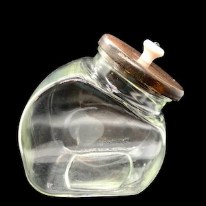 May include: A clear glass jar with a wooden lid and a white knob. The jar has a rounded shape and sits at an angle against a black background. The lid is dark brown with a light-colored knob.