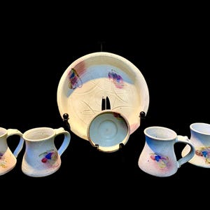 May include: A ceramic set consisting of four mugs and two plates, one large and one small. Each piece is off-white with a speckled texture and features abstract, colorful brushstroke accents in shades of blue, purple, and pink on the surface.