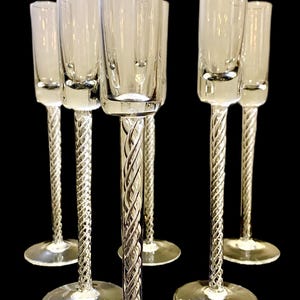 May include: Six clear champagne flutes with a tall, cylindrical bowl and a twisted stem design. The flutes have a wide, circular base and are arranged against a black background. The glasses are made of clear glass.