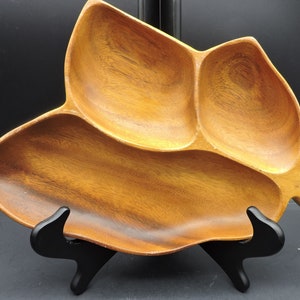 May include: A wooden leaf-shaped serving dish with three compartments. The dish is made of light brown wood and has a natural wood grain pattern. It is displayed on a black stand, showcasing its unique design. This decorative dish is ideal for serving snacks or appetizers.