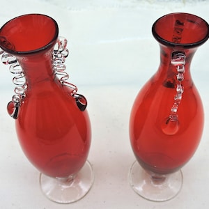May include: Two red glass vases with clear glass accents. One vase has a decorative handle, while the other has a clear glass chain. Both vases have a clear glass base and a wide opening.