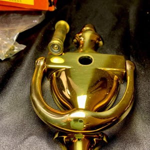 May include: A shiny brass door knocker with a classic design. The knocker features a curved handle and a decorative backplate. A small brass scope is also visible. The item is new and in its original packaging.