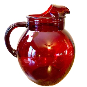 May include: A ruby red glass pitcher with a rounded body and a curved handle. The pitcher has a spout for pouring and reflects light, creating highlights. This vintage-style pitcher is suitable for serving beverages.
