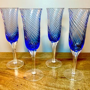 May include: Four blue-tinted champagne flutes with a spiral design. The flutes have a long, clear stem and a wide, circular base. The glasses are arranged on a wooden surface.