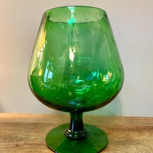May include: A large, green glass brandy snifter with a wide bowl and a short stem. The glass has a slight iridescent quality and is decorated with small, etched leaf designs around the rim. The snifter sits on a round base.