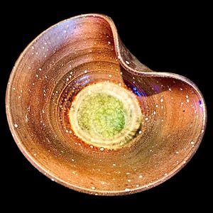 May include: A ceramic bowl with an irregular, organic rim shape. The exterior and upper interior feature a mottled, dark brown glaze with small white speckles. The center of the bowl has a circular, light green glazed area with a crackled texture.