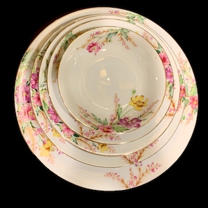 May include: A set of vintage white porcelain dishes with a floral design. The set includes plates and bowls of varying sizes, adorned with pink, yellow, and purple flowers, and gold trim. The arrangement is on a black background.