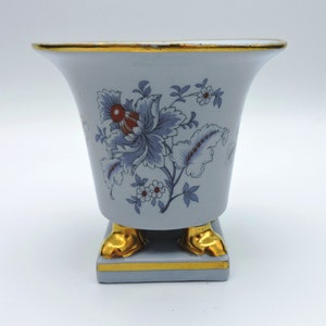 May include: A light blue ceramic vase with a flared top and gold trim. The vase features a floral design in blue, red, and white. The base has gold-colored feet.