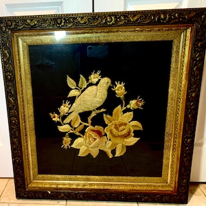 Peut inclure: Tableau encadré avec un oiseau brodé en or sur une branche ornée de roses dorées, sur fond noir. L'œuvre est encadrée dans un cadre en bois brun foncé orné avec des détails dorés, pour une esthétique vintage. Dimensions: environ 60 cm x 60 cm.