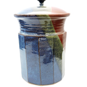 May include: A ceramic jar with a lid. The jar is a mix of blue, green, and red colors. The lid has a small knob on top. The jar has a geometric shape with vertical lines of color.