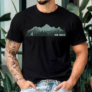 Könnte beinhalten: Schwarzes T-Shirt mit einem Berg- und Läufer-Motiv. Das Design zeigt eine Läufer-Silhouette, Bäume und Berge in Grün- und Grautönen mit dem Text "RUN TRAILS" in Weiß.
