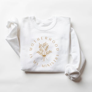 May include: White long-sleeve sweatshirt with a gold graphic print that reads "Motherhood is my ministry" with a floral design.