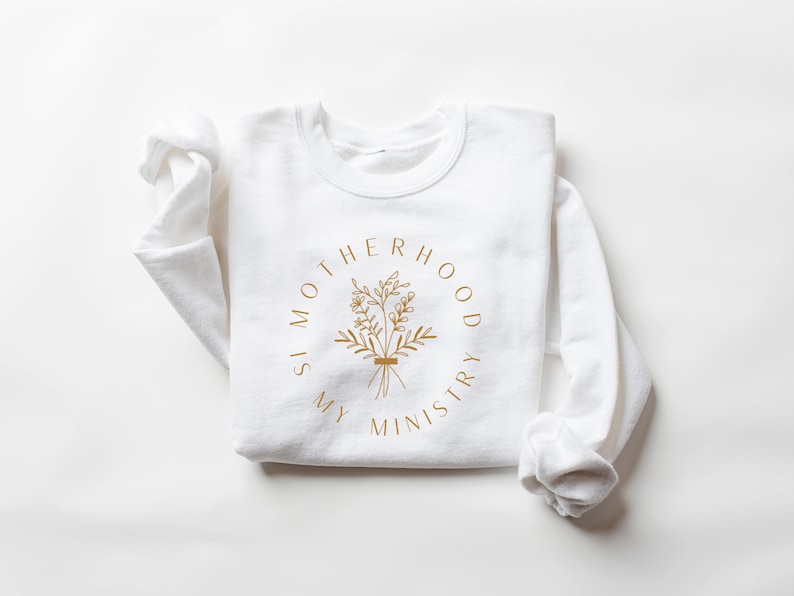 May include: White long-sleeve sweatshirt with a gold graphic print that reads "Motherhood is my ministry" with a floral design.
