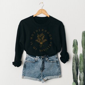 May include: Black sweatshirt with a gold graphic that says "Motherhood is my ministry" with a floral design. The sweatshirt is hanging on a hanger and is paired with denim shorts.