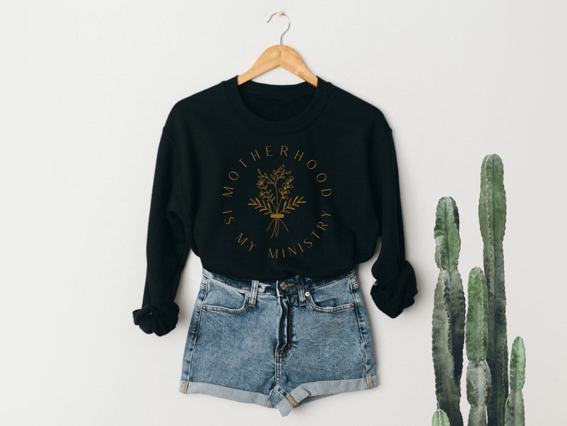 May include: Black sweatshirt with a gold graphic that says "Motherhood is my ministry" with a floral design. The sweatshirt is hanging on a hanger and is paired with denim shorts.