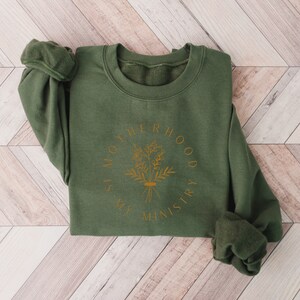 May include: A green sweatshirt with a gold graphic that says "Motherhood is my ministry" with a floral design.