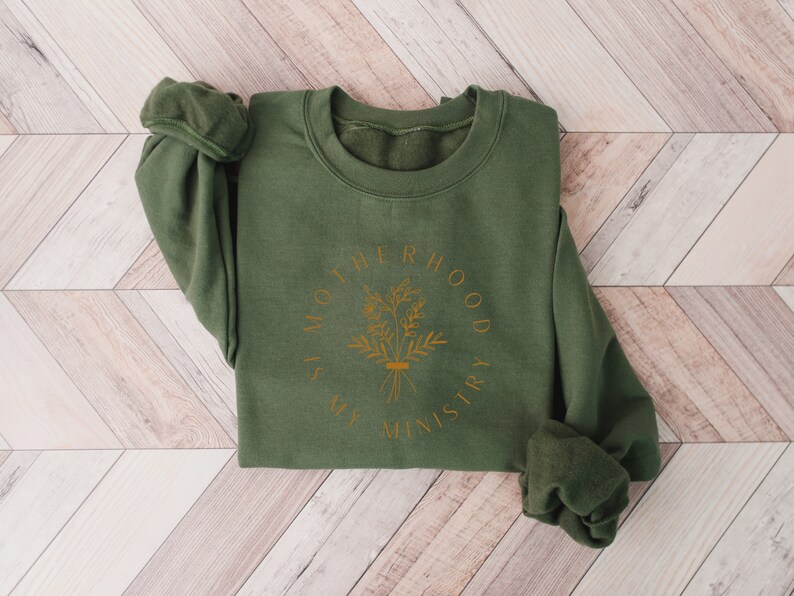 May include: A green sweatshirt with a gold graphic that says "Motherhood is my ministry" with a floral design.