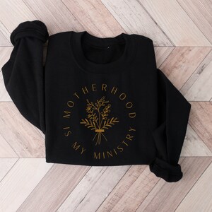 May include: Black sweatshirt with a gold graphic that reads "Motherhood is my ministry" with a floral design.