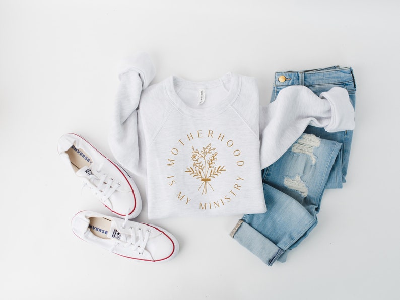 May include: A gray sweatshirt with the text "Motherhood is my ministry" in a gold circle with a floral design. The sweatshirt is laid out on a white surface with a pair of white sneakers and blue jeans.