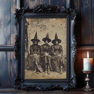 Puede incluir: Una imagen enmarcada en negro de tres mujeres con sombreros de bruja y vestidos. La imagen está sobre una superficie de madera oscura con una vela encendida y una calabaza tallada.