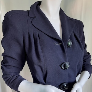 May include: A navy blue blazer with a tailored fit. The jacket features a notched lapel, three large black buttons, and a silver bar closure. The sleeves are three-quarter length.