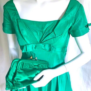 May include: A vibrant emerald green dress with a square neckline, short sleeves featuring small bows, and a pleated bodice with a central bow at the waist. A matching emerald green clutch bag with a bow detail and a metallic clasp is held in front.