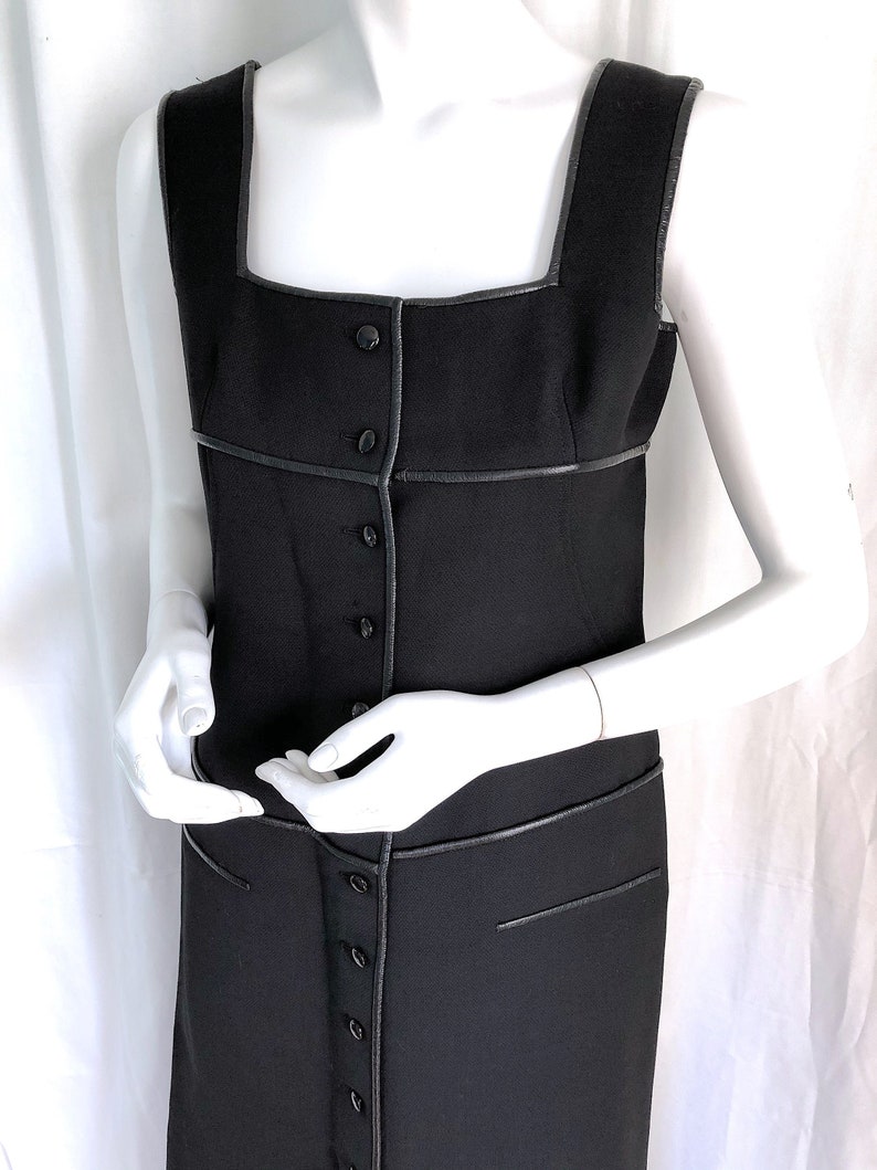 May include: A black sleeveless dress with a square neckline and a row of buttons down the front. The dress features black trim along the neckline, armholes, and seams. The dress is displayed on a mannequin.