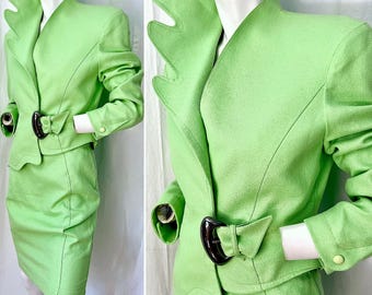 May include: A lime green two-piece suit consisting of a jacket and skirt. The jacket features a high, scalloped collar, long sleeves with single buttons, and a large, dark brown asymmetrical buckle closure at the waist. The skirt is knee-length.