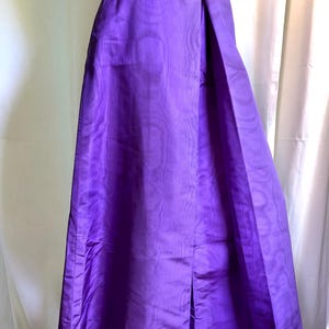 May include: A floor-length purple skirt featuring a subtle, tonal moiré pattern throughout the fabric. The skirt has a high waistband and a straight, structured silhouette with a prominent vertical front slit that extends from the hem upward.