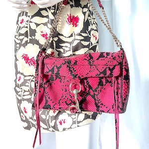 May include: A pink and black snakeskin-patterned shoulder bag with a silver chain strap. The bag has a rectangular shape with a flap closure and long, thin tassels. The bag is held by a mannequin wearing a floral dress.