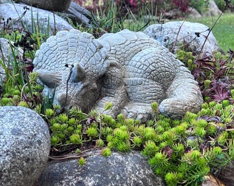 Puede incluir: Una escultura de hormigón gris de un dinosaurio durmiendo, un Triceratops, tumbado en un lecho de musgo verde y rocas.