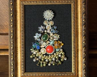 May include: A framed artwork featuring a Christmas tree made of various vintage jewelry pieces, including rhinestones, pearls, and colorful stones. The tree is set against a black fabric background and framed in a gold ornate frame.