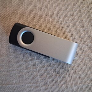 Könnte beinhalten: Ein silberner und schwarzer USB-Stick. Der Stick hat eine gebürstete Metalloberfläche am Hauptkörper und ein schwarzes Ende mit einem kreisförmigen Knopf. Am Ende ist eine kleine Metallschlaufe angebracht.