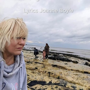Puede incluir: Una mujer con una sudadera gris mira hacia el mar. El texto "Dancing with Memories Lyrics Joanne Boyle" está en la parte superior. Una familia camina por la orilla rocosa con un perro bajo un cielo nublado.