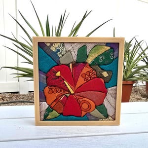 May include: A square framed textile art piece featuring a central hibiscus flower constructed from various fabric scraps in shades of red, orange, and yellow. The background consists of geometric fabric patches in teal, purple, and cream patterns.