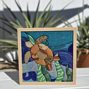 May include: A square fabric art piece featuring a sea turtle made of various patterned orange and brown fabric patches. The turtle swims against a background of blue and teal fabric waves and green seaweed shapes, all enclosed in a light wood frame.