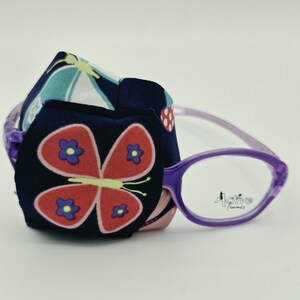 May include: A pair of purple spectacles with a dark blue fabric eye patch attached to one lens. The eye patch features a large red butterfly graphic with blue flower centres. The text Active frames is printed on the clear spectacle lens.