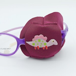 May include: Purple eye patch with an adjustable strap and a dinosaur applique. The patch is made of soft fabric and is designed to be comfortable for children. The dinosaur applique is pink, green, and red with white polka dots.