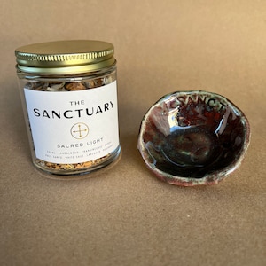Könnte beinhalten: Ein Glasgefäß mit goldenem Deckel, gefüllt mit getrockneten Kräutern und mit "The Sanctuary Sacred Light Copal, Sandalwood, Frankincense, Myrrh, Palo Santo, White Sage, Lavender, Rosemary" beschriftet. Eine kleine, braune Keramikschale mit grüner Glasur.