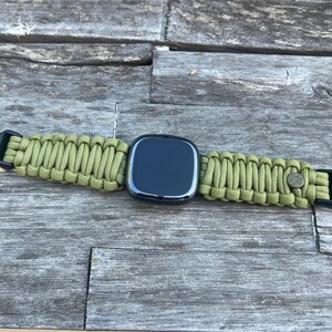 May include: A black smartwatch with a green paracord strap. The strap has a buckle on one end and a metal clasp on the other.