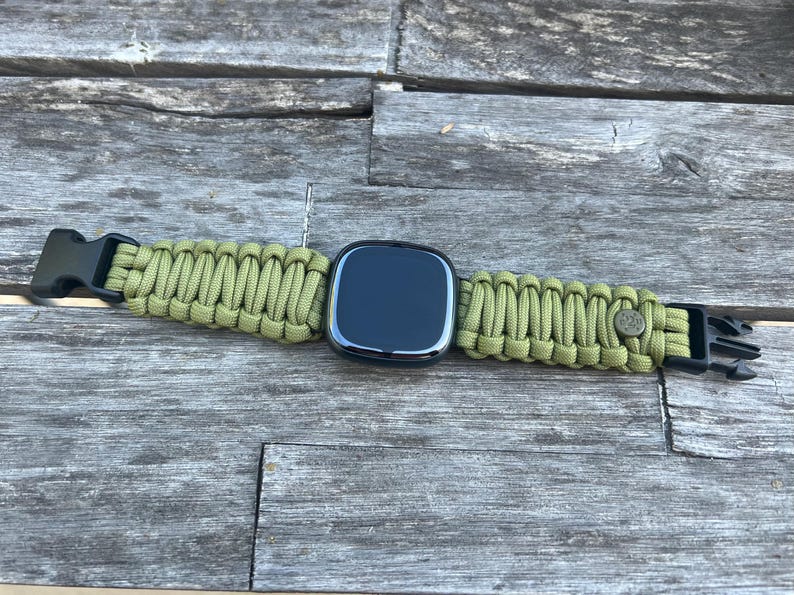 May include: A black smartwatch with a green paracord strap. The strap has a buckle on one end and a metal clasp on the other.