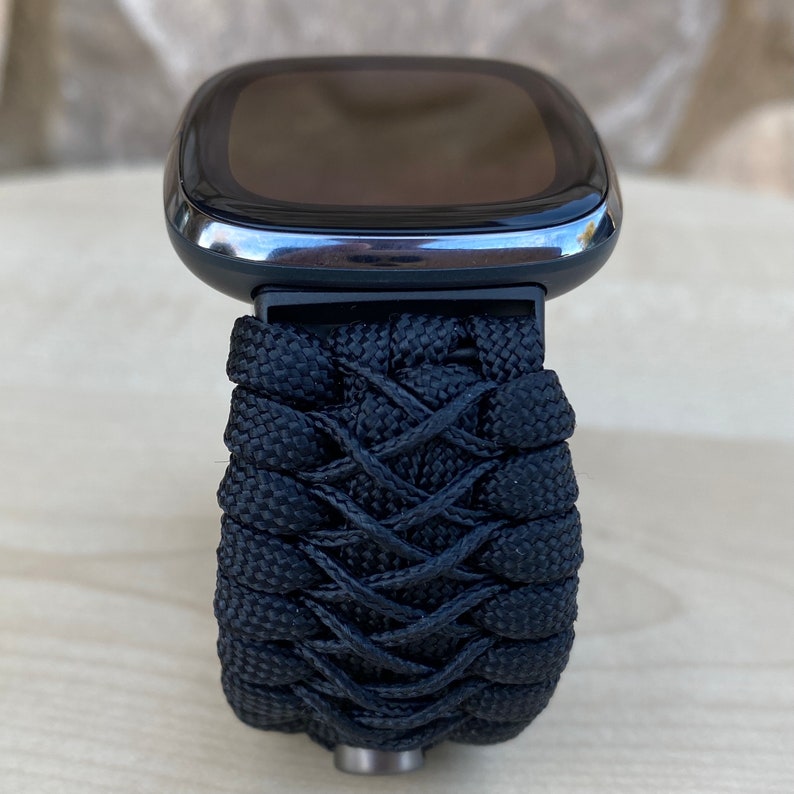 May include: A black smartwatch with a black woven paracord strap. The watch face is black and has a silver bezel.