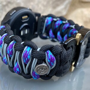 May include: A watch band crafted from black, purple, and blue paracord featuring a woven design. The band includes a metallic silver bead with star engravings and a black metal buckle with a gold-toned release mechanism attached to a black watch.