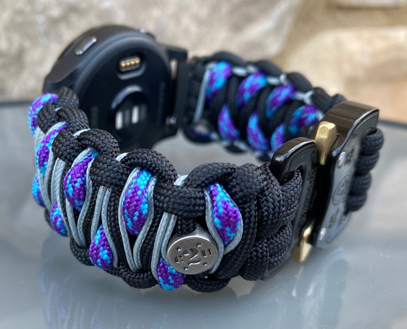 May include: A watch band crafted from black, purple, and blue paracord featuring a woven design. The band includes a metallic silver bead with star engravings and a black metal buckle with a gold-toned release mechanism attached to a black watch.
