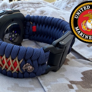 Op de afbeelding: Een zwarte smartwatch met een marineblauwe en bruine paracord band. De band heeft een rode accentkleur en een zwarte sluiting. Een embleem van het United States Marine Corps is zichtbaar op de achtergrond.