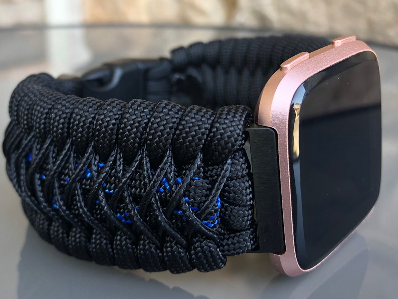 Puede incluir: Una correa de reloj de paracord negra con detalles azules, unida a un smartwatch de color rosa dorado con una pantalla negra.