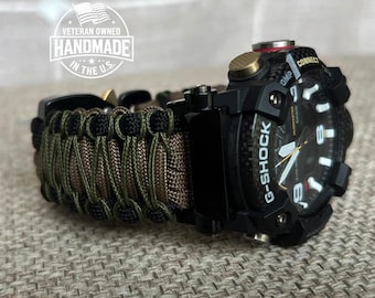 Puede incluir: Un reloj G-Shock negro con una correa de paracord verde y negro. La esfera del reloj tiene un fondo blanco con marcas negras y una aguja de segundos roja. El reloj tiene un botón negro en la parte superior derecha y un botón dorado en la parte superior izquierda. La correa está hecha de paracord y es ajustable. El reloj es una opción popular para los entusiastas de las actividades al aire libre y aquellos que necesitan un reloj duradero y confiable.